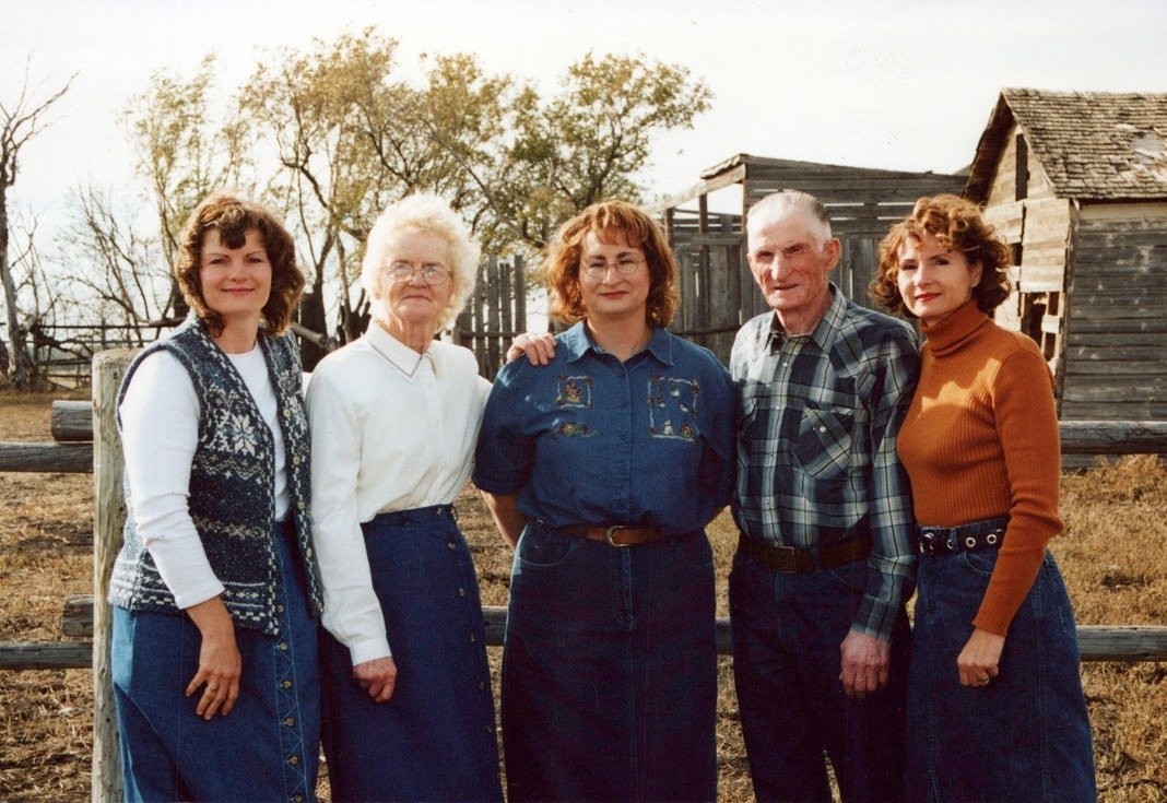 Three generations of a family standing together by a fence on a prairie farm, with old wooden barns behind them